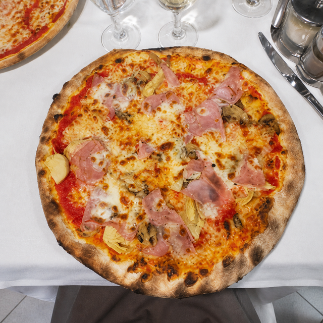 Pizza rotonda con prosciutto e funghi su una tovaglia bianca, vista dall'alto.