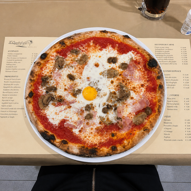 Una grande pizza su un piatto con salsa di pomodoro, formaggio fuso, funghi e un uovo crudo sopra, su un tavolo di ristorante.