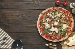 Tavolo di legno con una pizza condita con basilico, formaggio e pomodori, oltre a olio d'oliva e altri ingredienti nelle vicinanze.