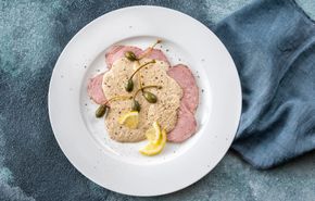 Pesce gourmet impiattato con capperi, salsa rosa e limone su un piatto bianco sopra una tovaglia blu.