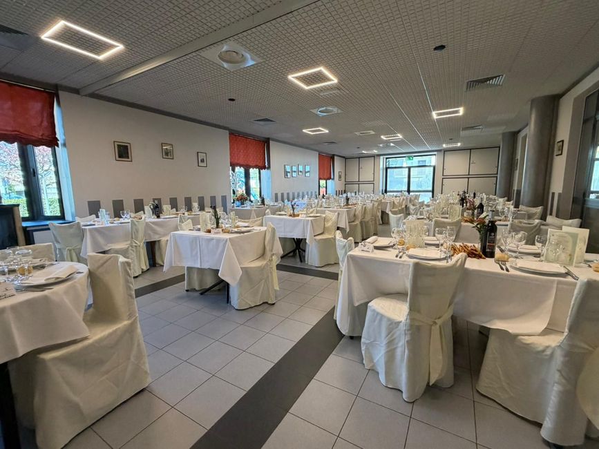 Sala da banchetto vuota con tavoli rotondi, tovaglie bianche e tende rosse alle finestre.