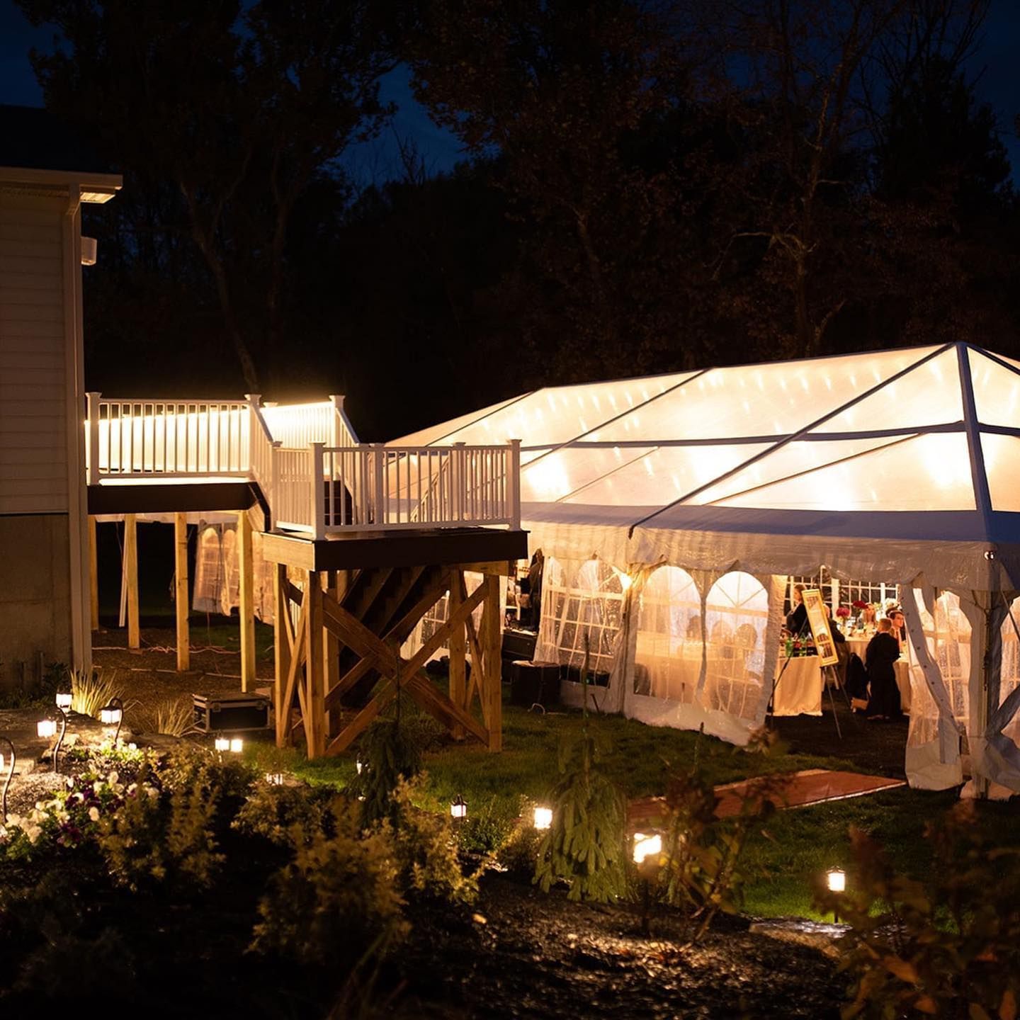 A backyard party at night: a lit clear tent, a wooden deck, and garden lights.