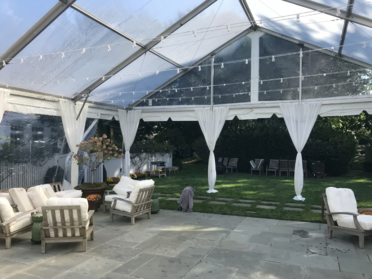 Clear tented outdoor lounge with white chairs, sheer drapes, and stone patio beside a grassy yard