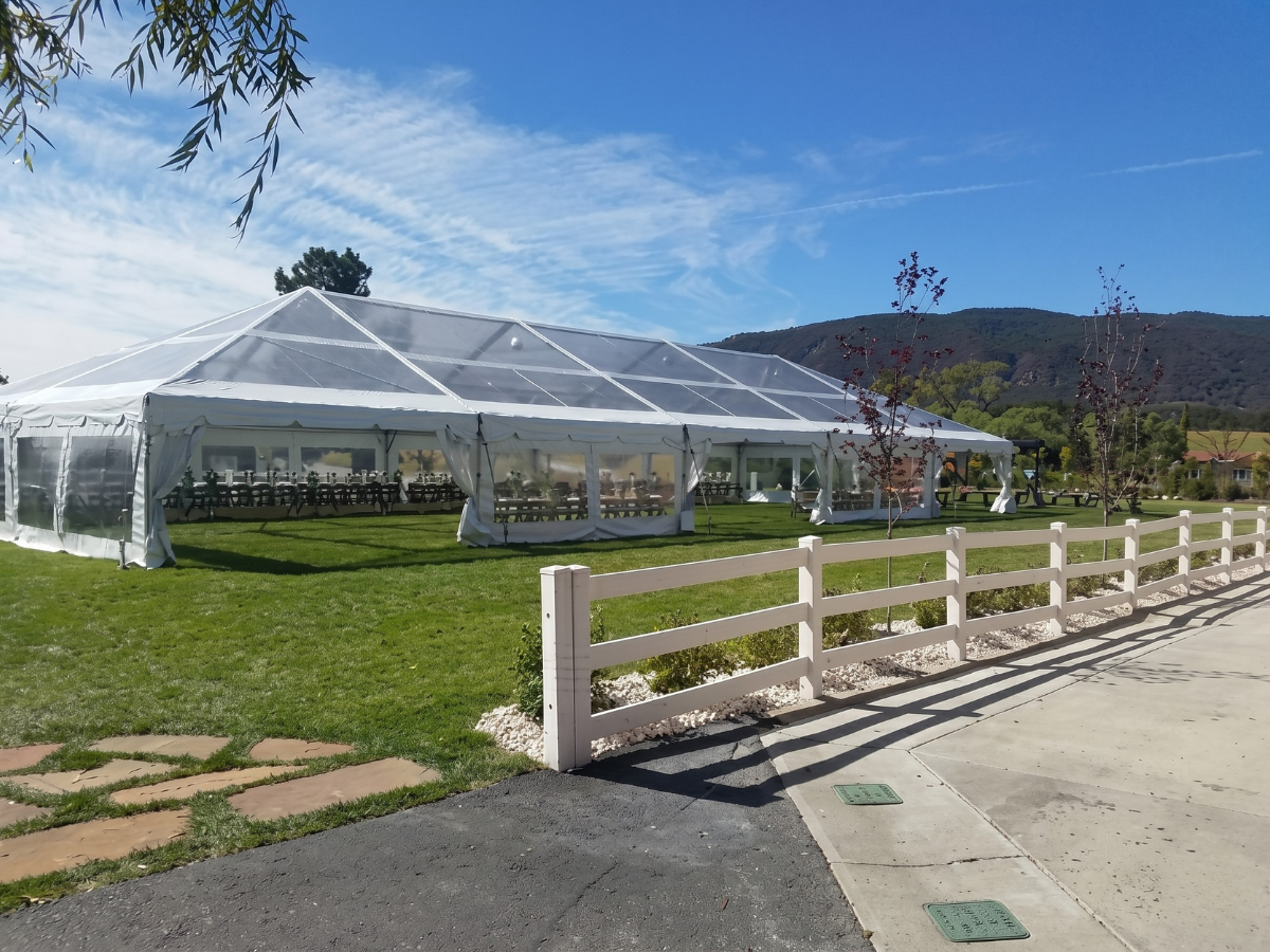 White event tent on a grassy lawn beside a road, with mountains and blue sky in the background.