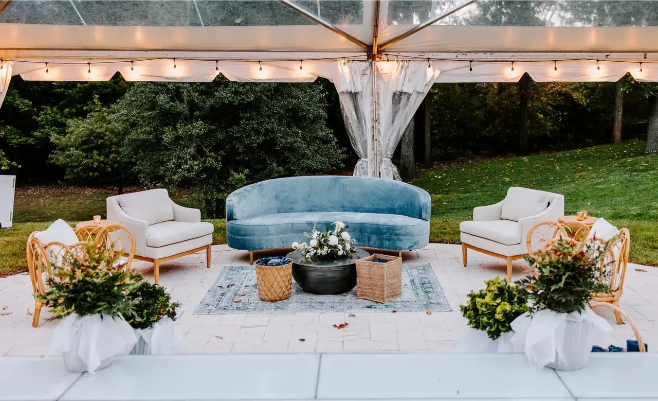 Outdoor lounge area with blue sofa, two cream armchairs, coffee table, and greenery under a clear tent.