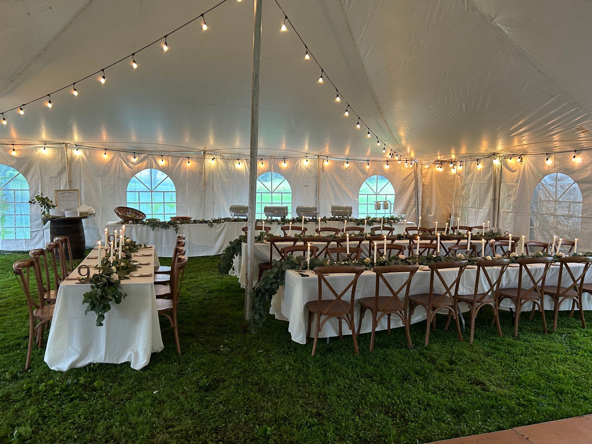 Wedding reception tent set up with tables, chairs, and string lights, on a grassy area.