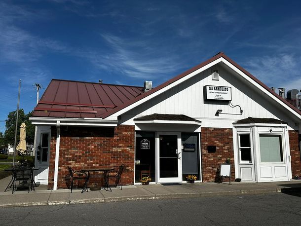 Small brick-and-white building with a red metal roof and a sign above the entrance on a sunny day