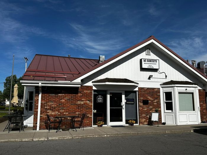 Small brick-and-white building with a red metal roof and a sign above the entrance on a sunny day