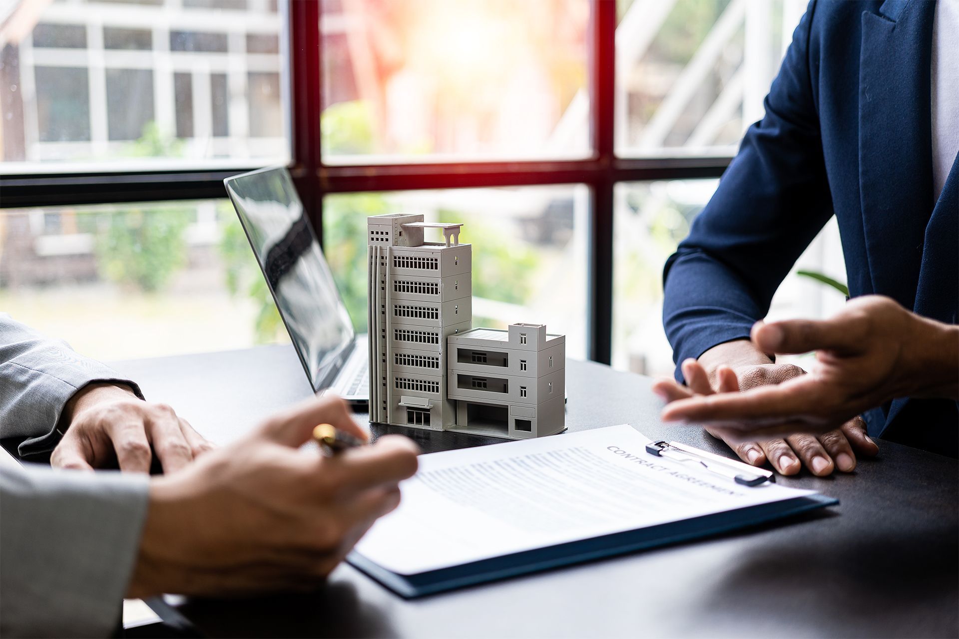 Two people reviewing real estate documents with a model building and laptop on the table.