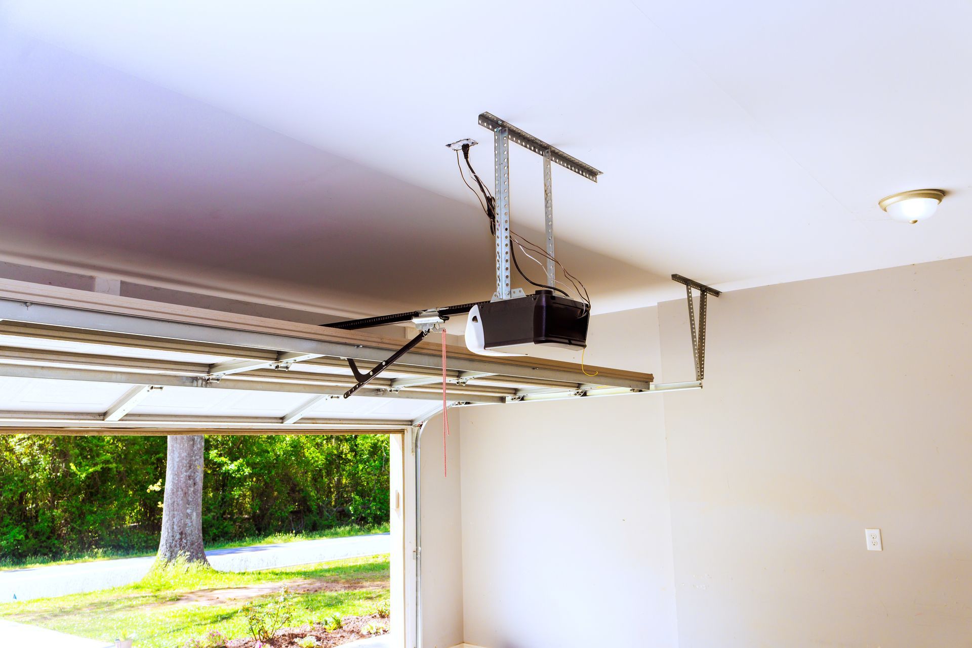 Automatic garage door opener motor mounted on ceiling track with open door view outside.