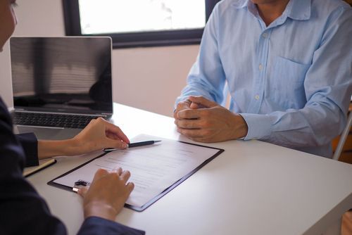 A person in a business suit points to a document on a clipboard held by an individual in a light blue button-down shirt.