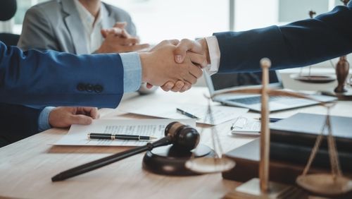 Two people in business suits shake hands over a legal document, with a wooden gavel and balance scales on the desk.
