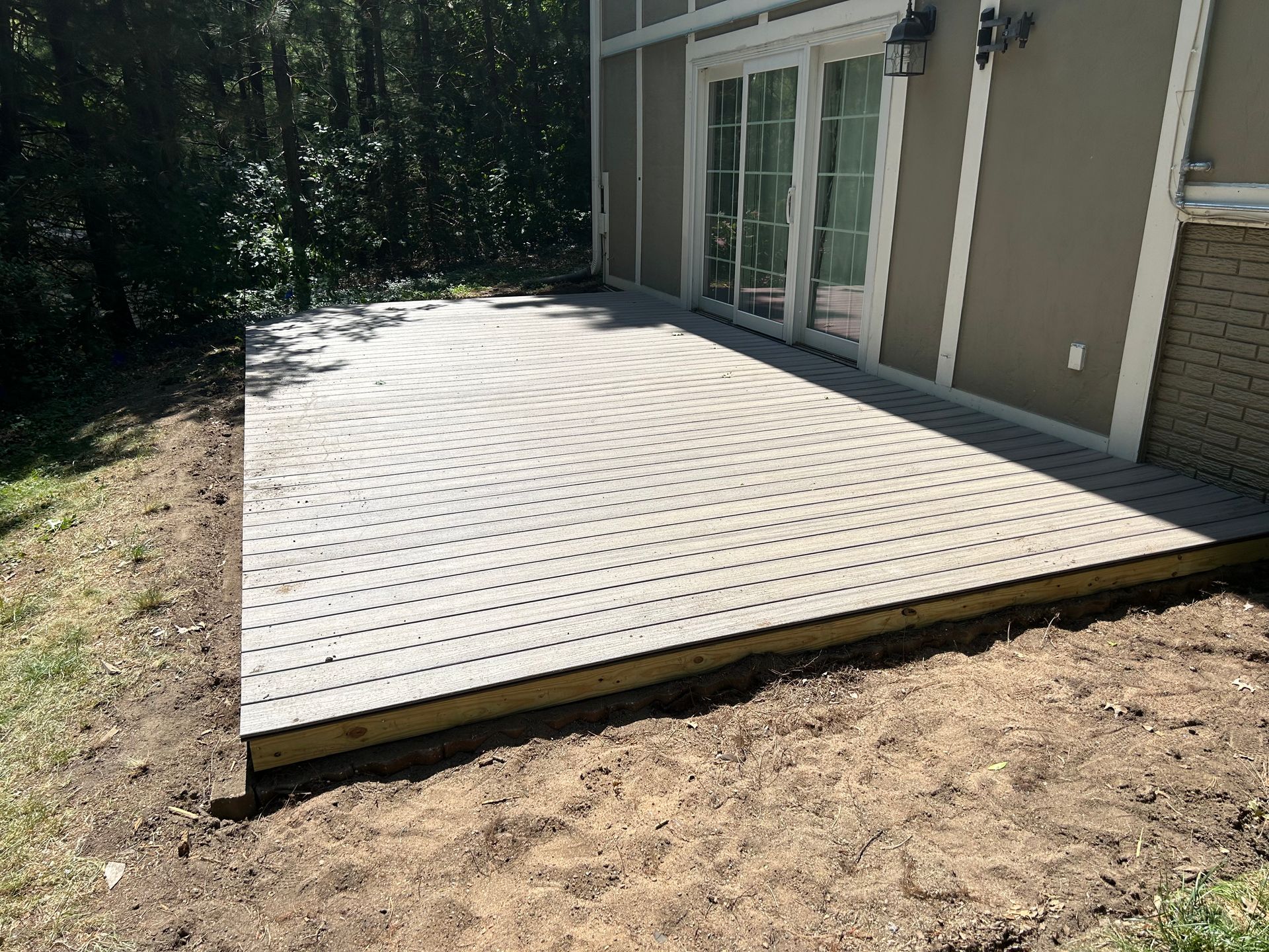 Raised wooden deck patio beside a house, with sliding glass doors and a wooded yard nearby.
