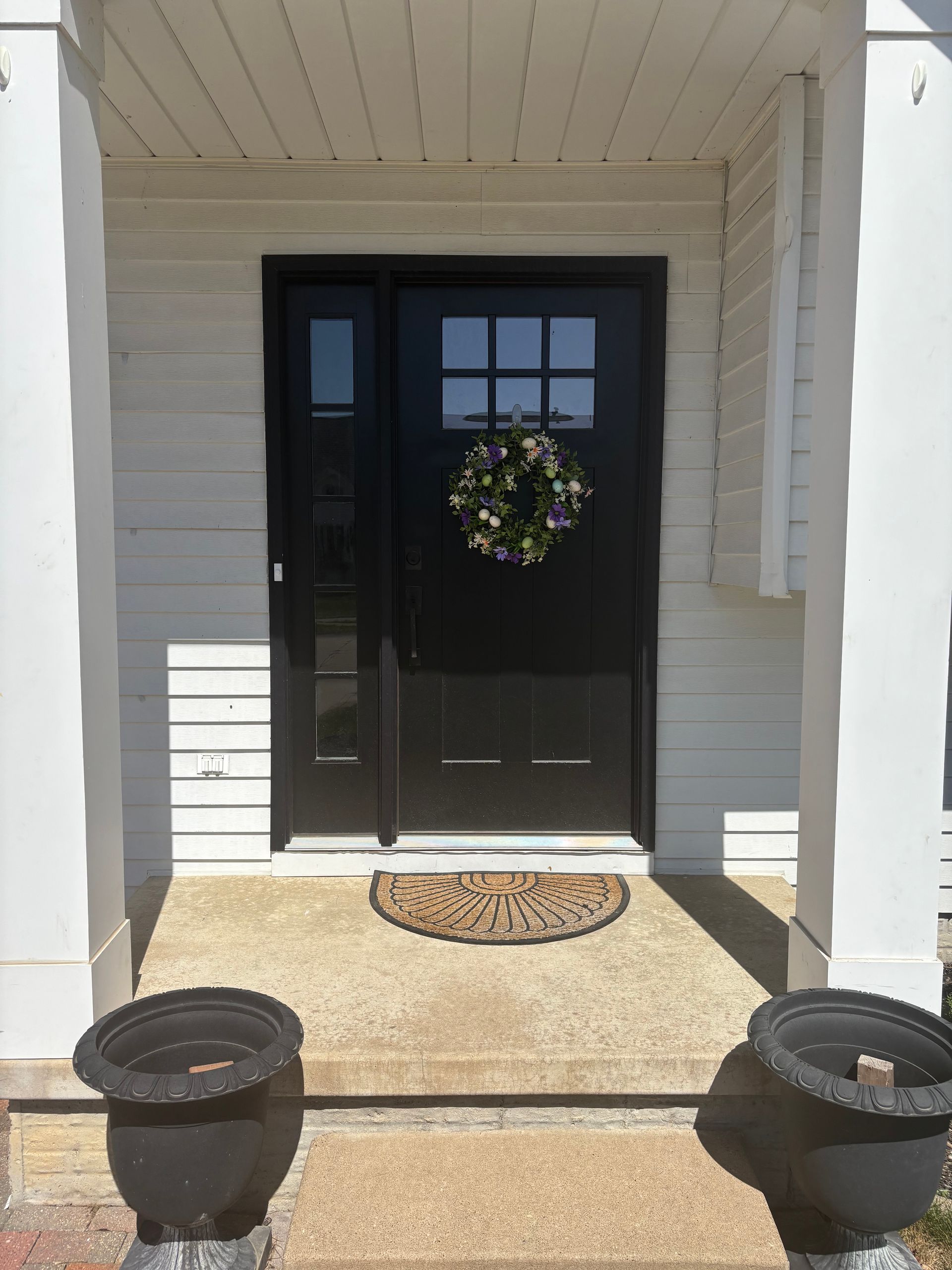 Front porch with black front door, wreath, doormat, and two planters in bright sunlight
