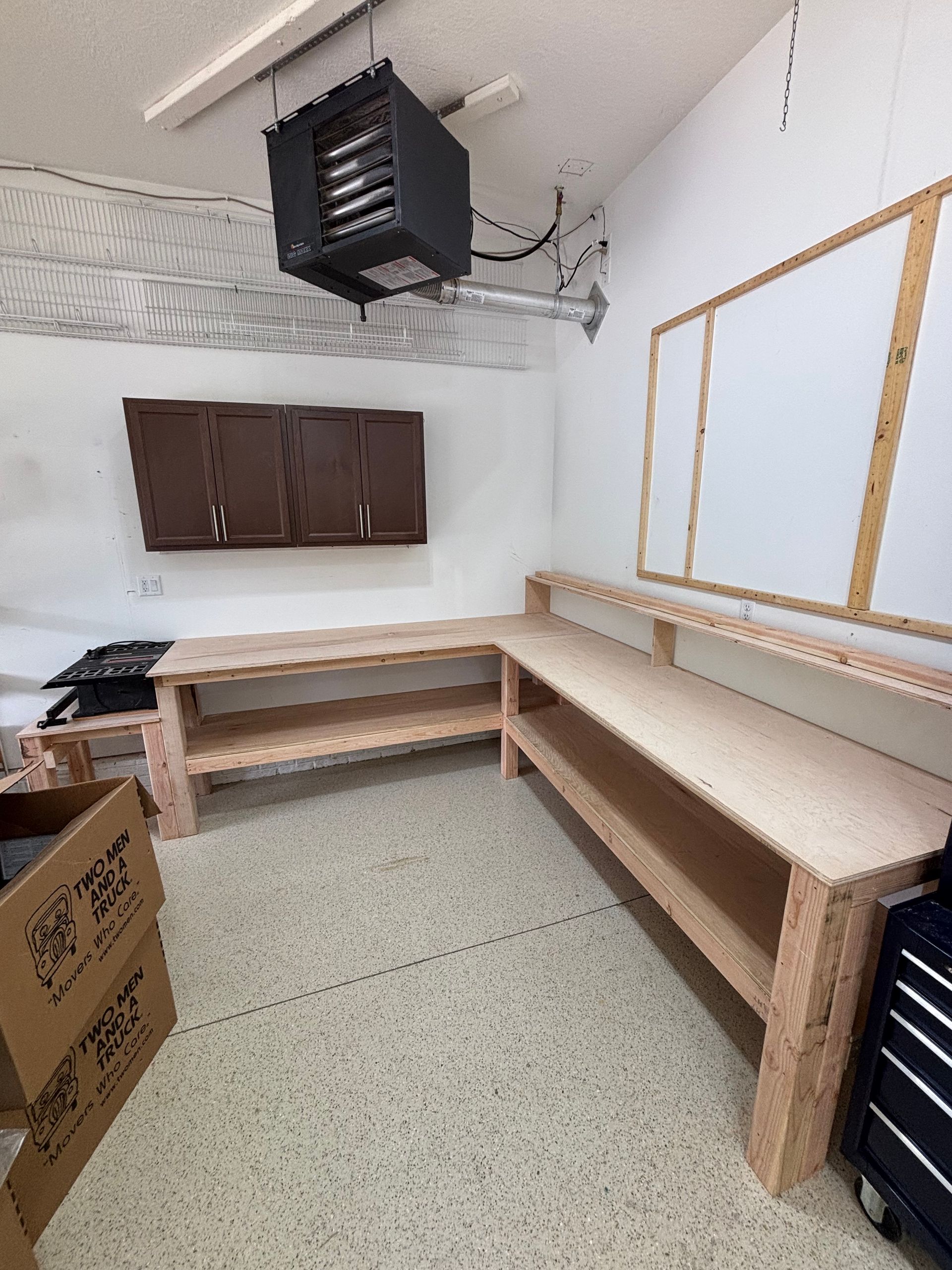 Empty workshop with L-shaped wooden workbench, wall cabinets, and overhead vent unit.