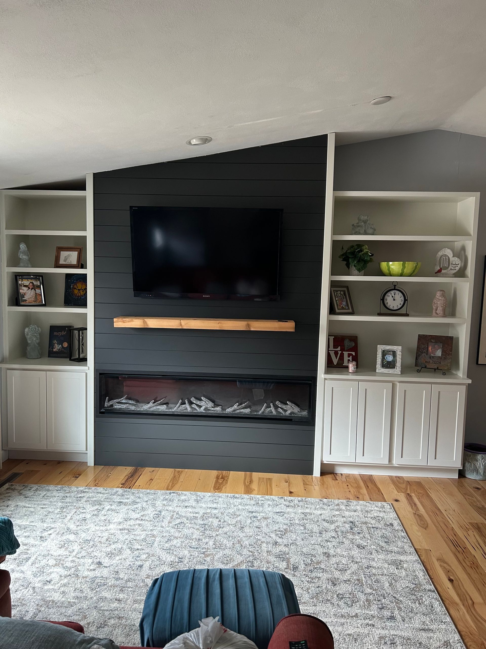 Living room wall with built-in shelves, large TV, fireplace, and gray rug over hardwood floor