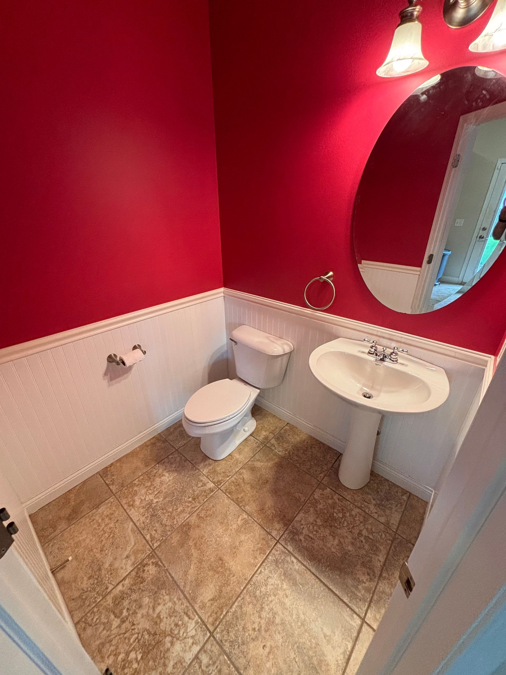 Small bathroom with red walls, white wainscoting, pedestal sink, toilet, round mirror, and tan tile floor.