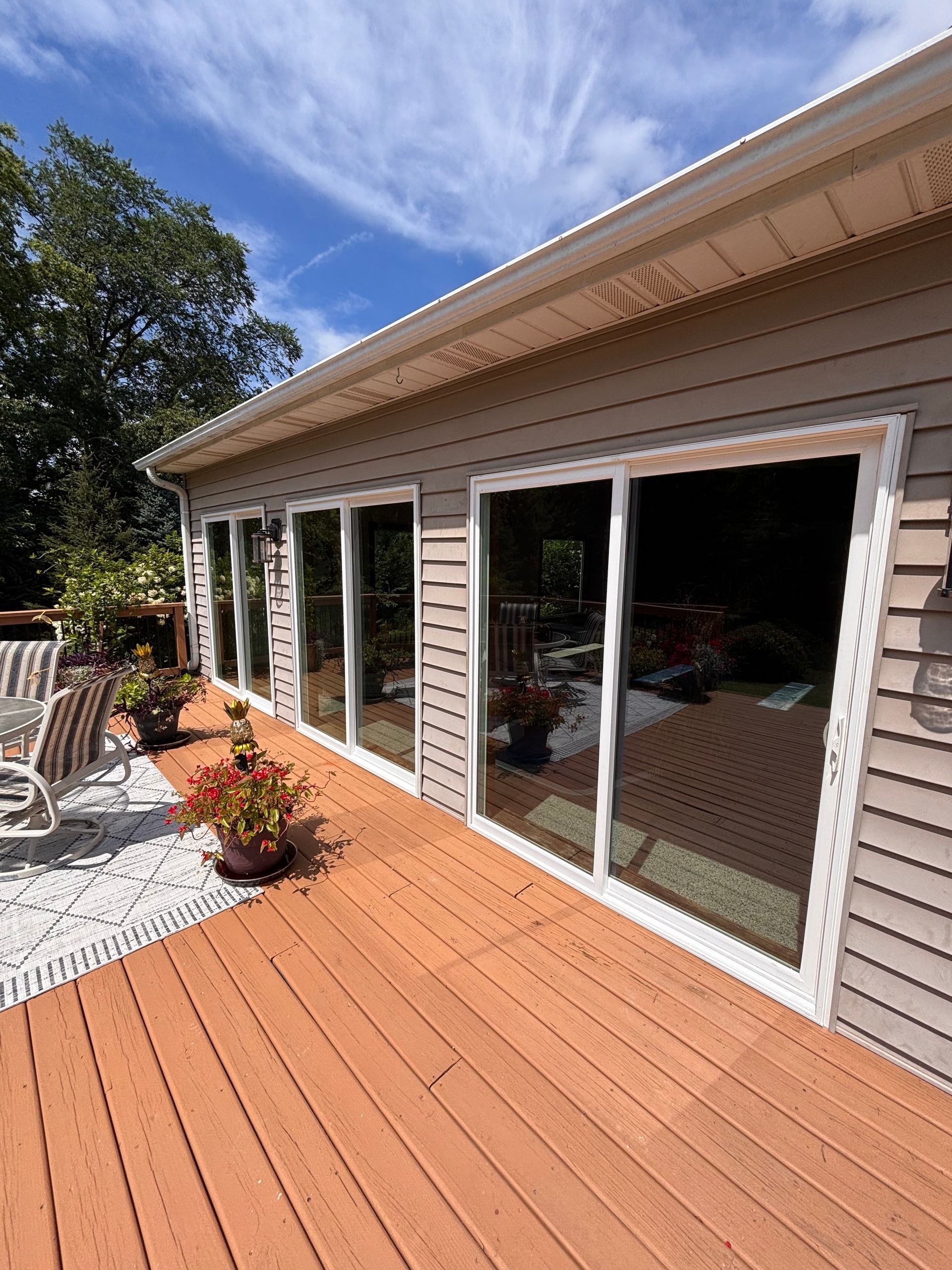 Wooden deck beside a house with large sliding glass doors and potted plants in bright sunlight