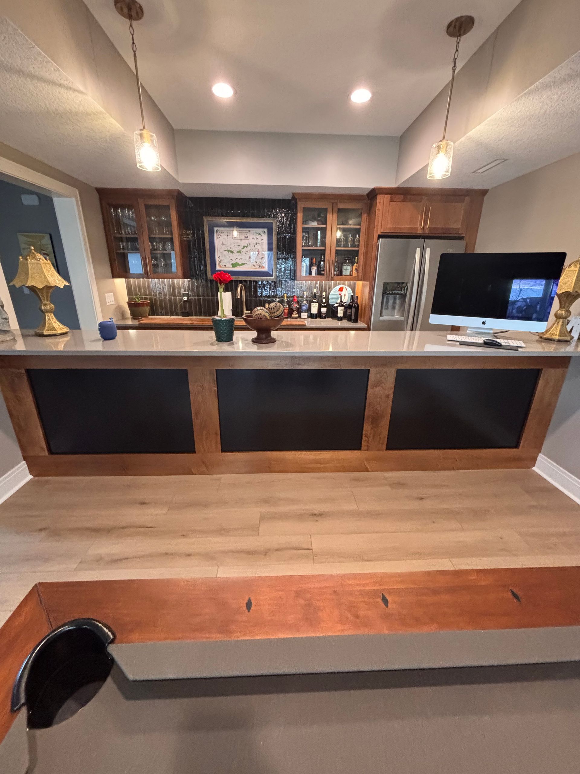 Modern home bar with wood counter, black panels, shelves, pendant lights, and TV in the background