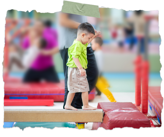 Child balancing on a gym foam beam, with blurred children and colorful play equipment in the background