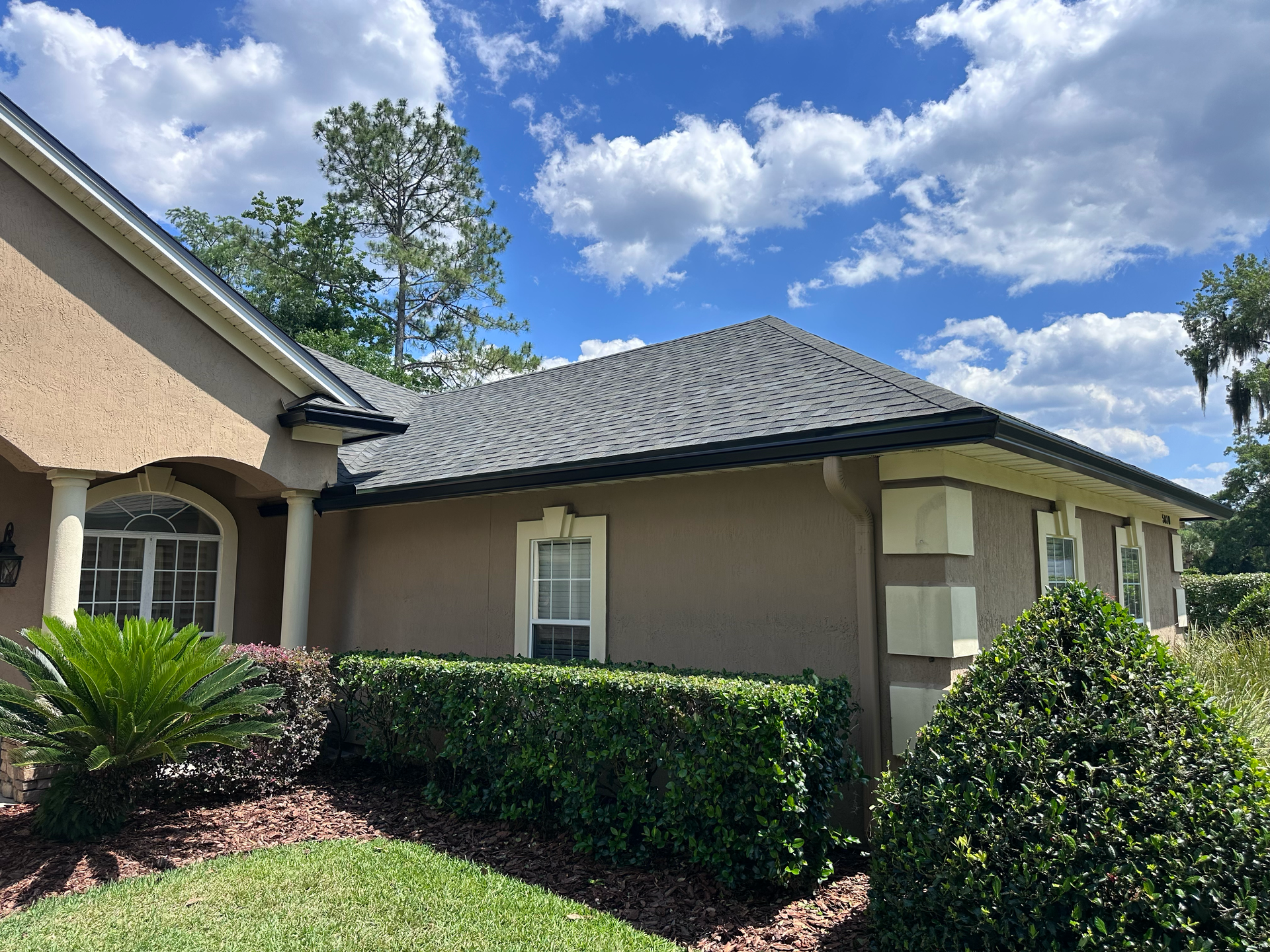Suburban house exterior with beige walls, gray roof, manicured hedges, and blue sky with clouds