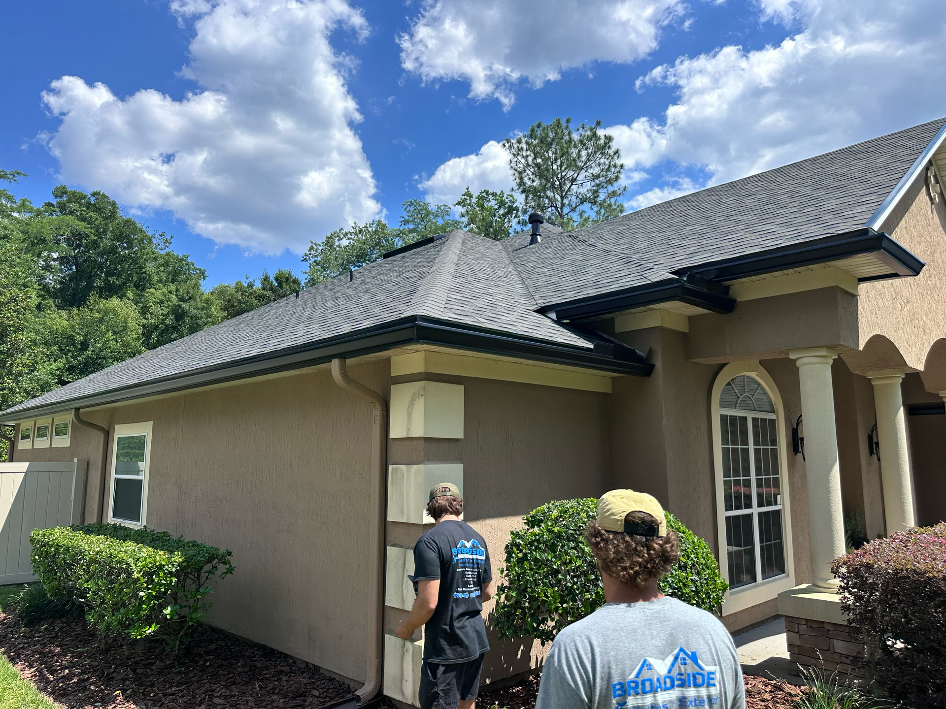 Two people inspecting the side of a beige house with a gray roof on a sunny day