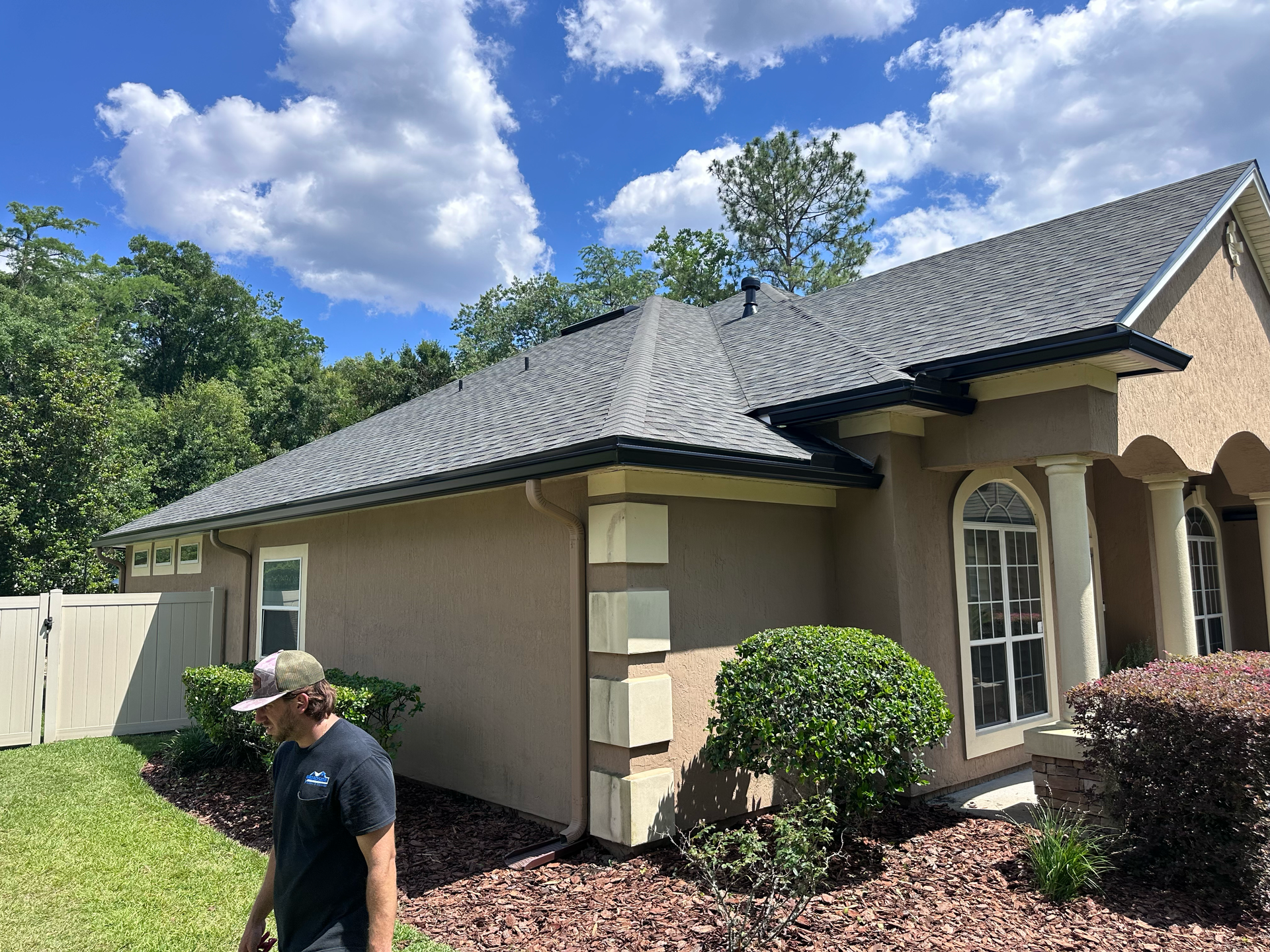 Man standing beside a beige house with a gray roof, landscaped yard, and blue sky with clouds