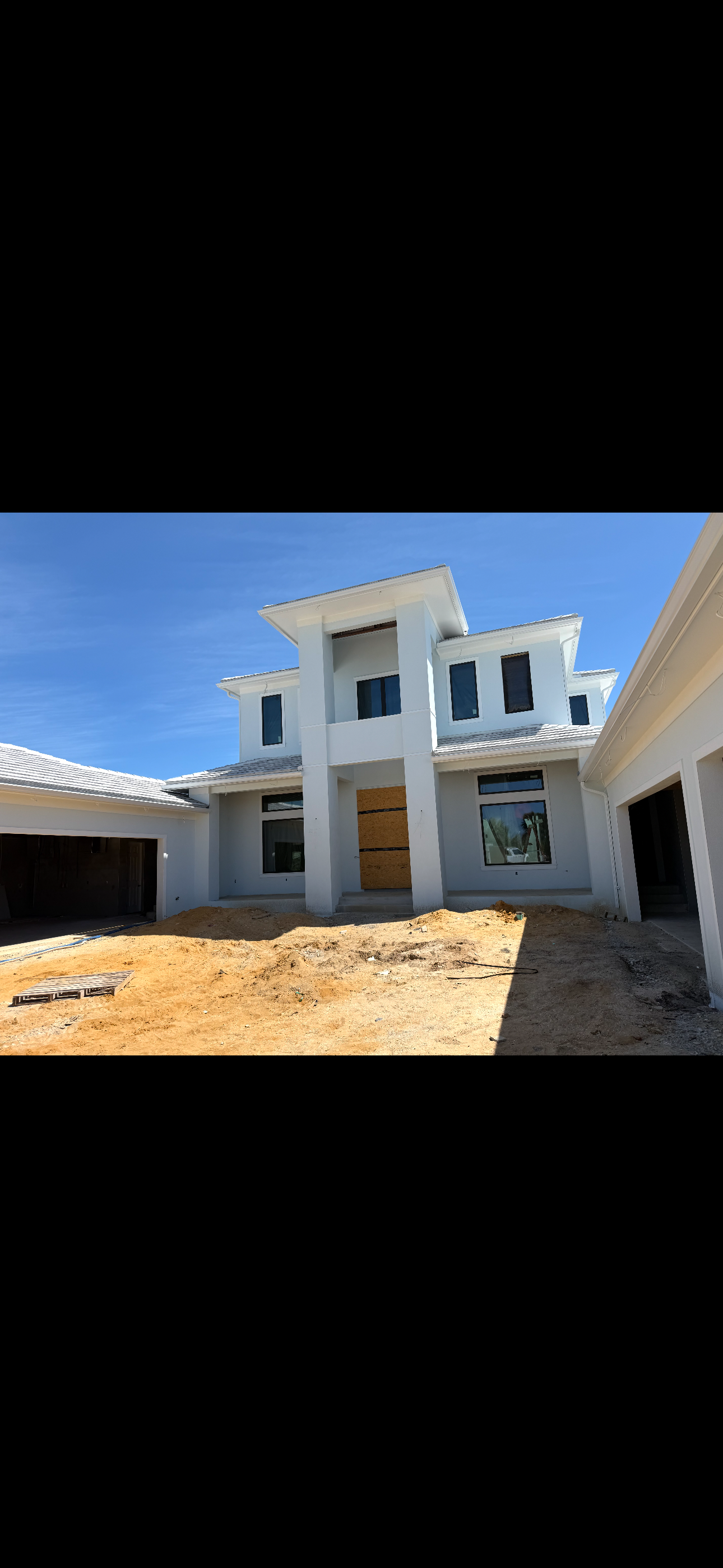 Modern two-story white house under construction with garage and unfinished dirt driveway in bright sunlight