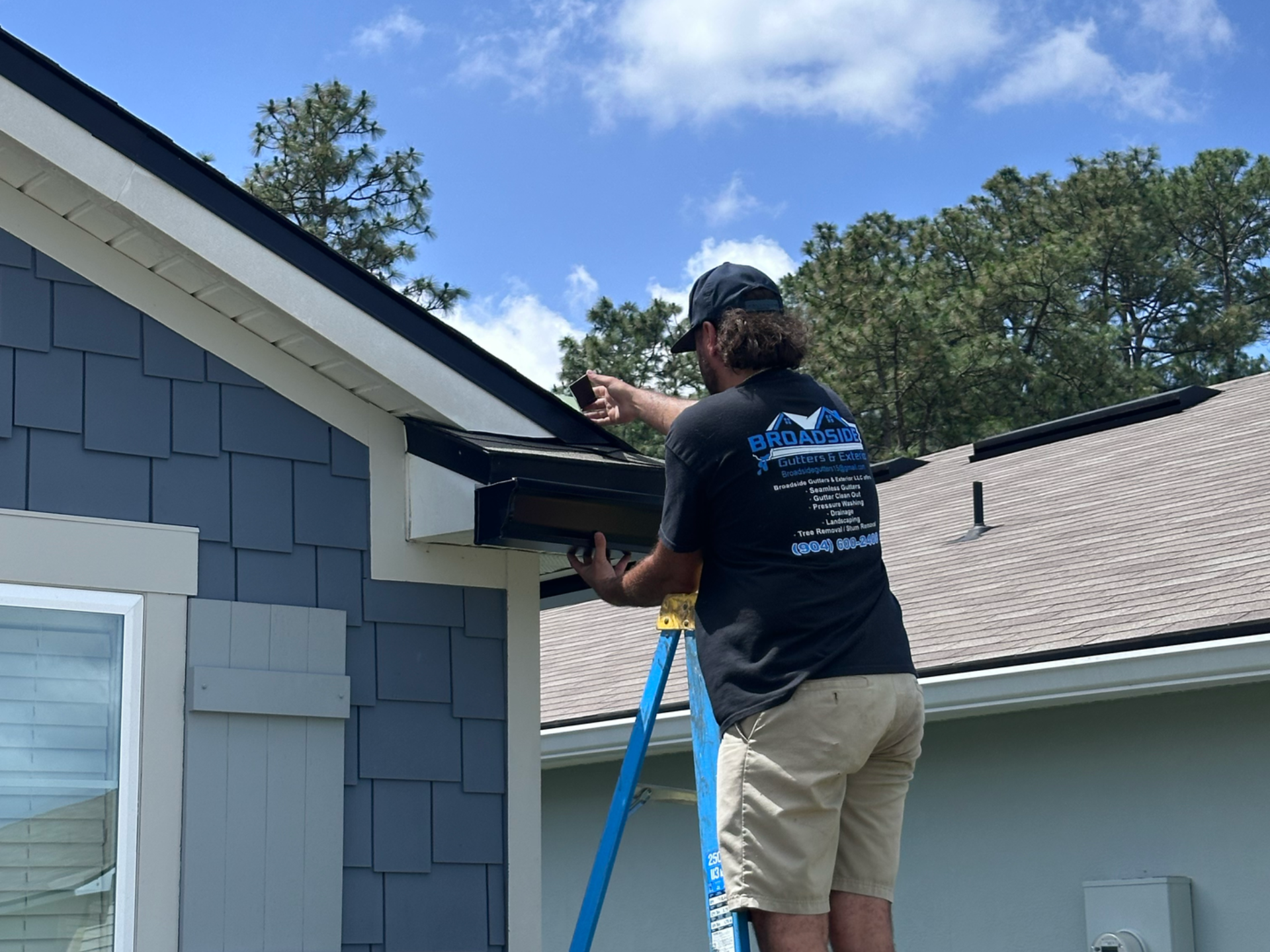 Worker on ladder repairing blue house roof eave beside a light gray house.