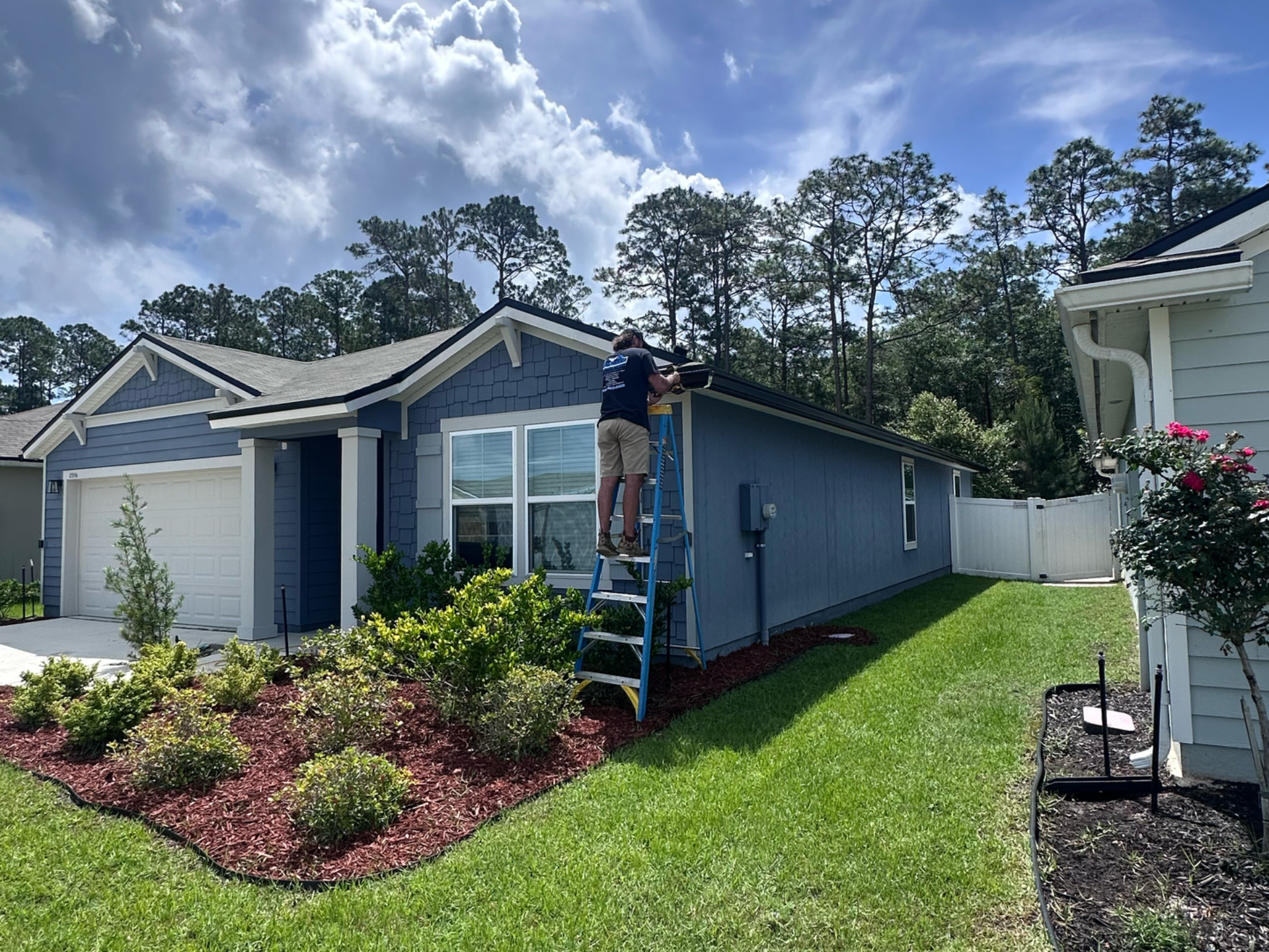 Worker painting the side of a blue house on a ladder in a sunny suburban yard