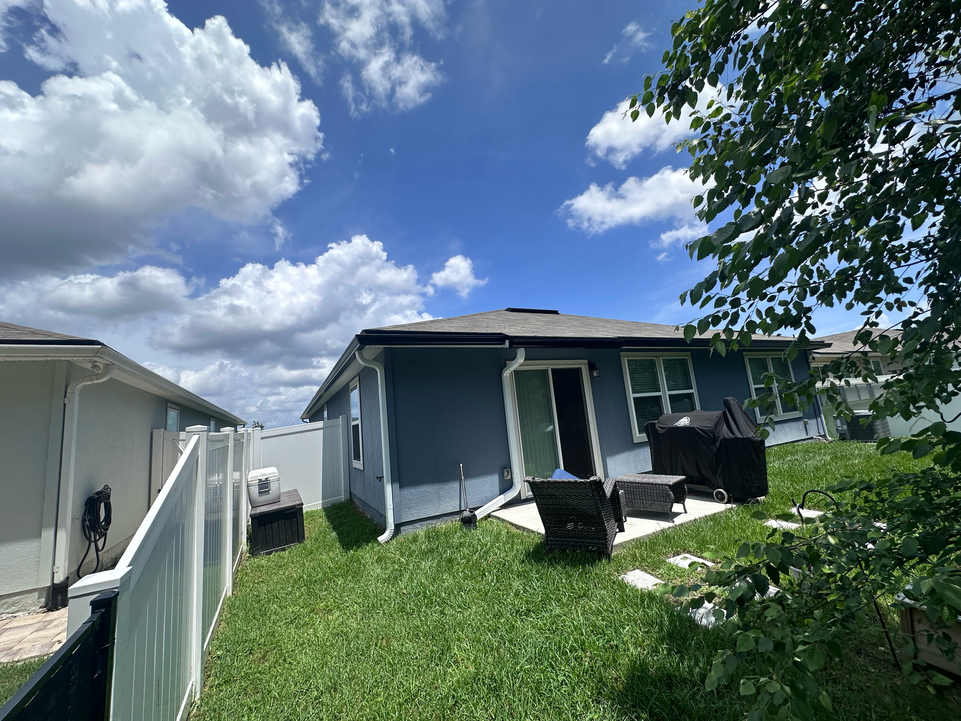 Backyard with patio chairs, white fence, and gray house under a partly cloudy sky
