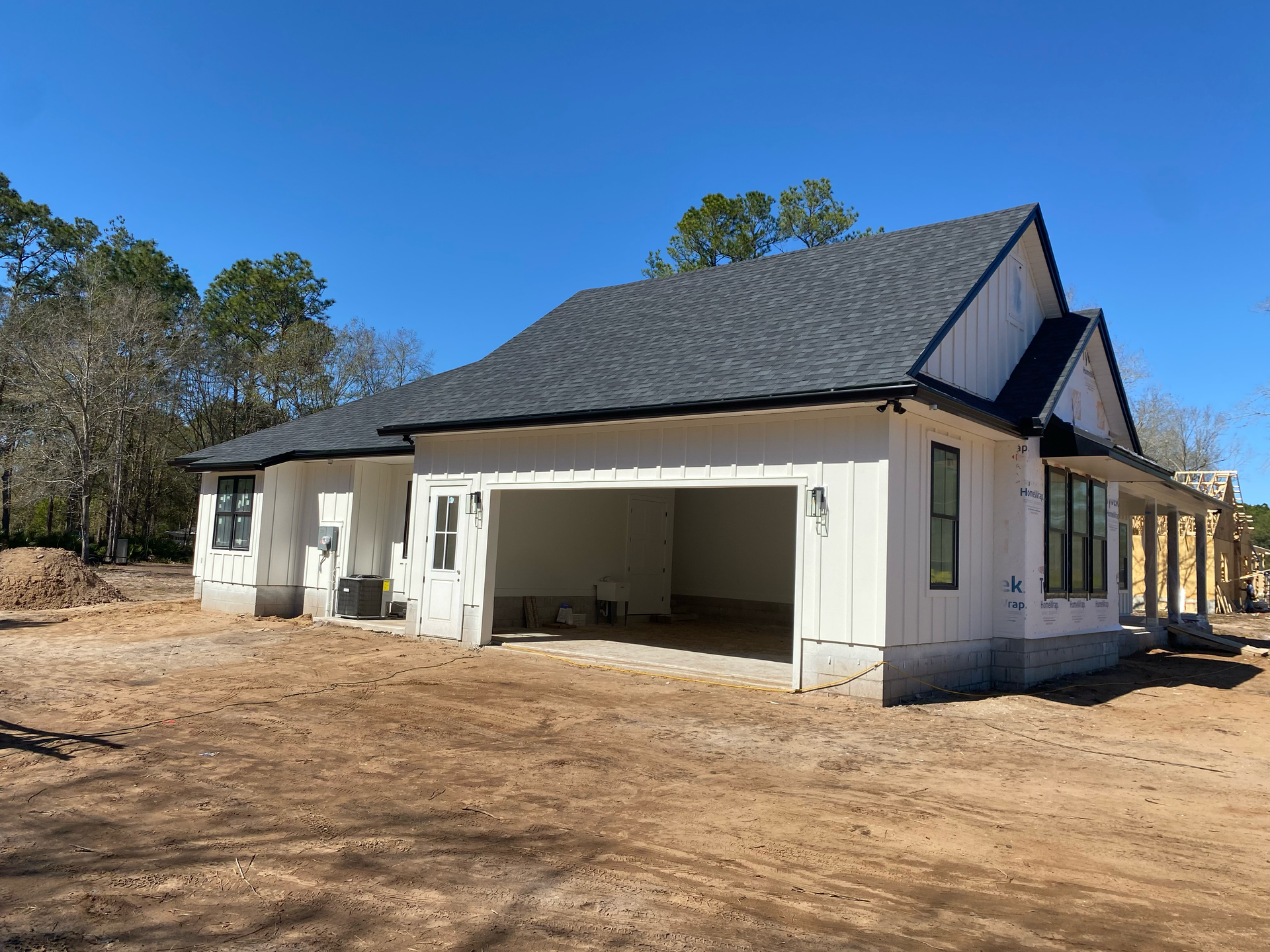 White house under construction with open garage, dark roof, and bare dirt yard under a clear blue sky