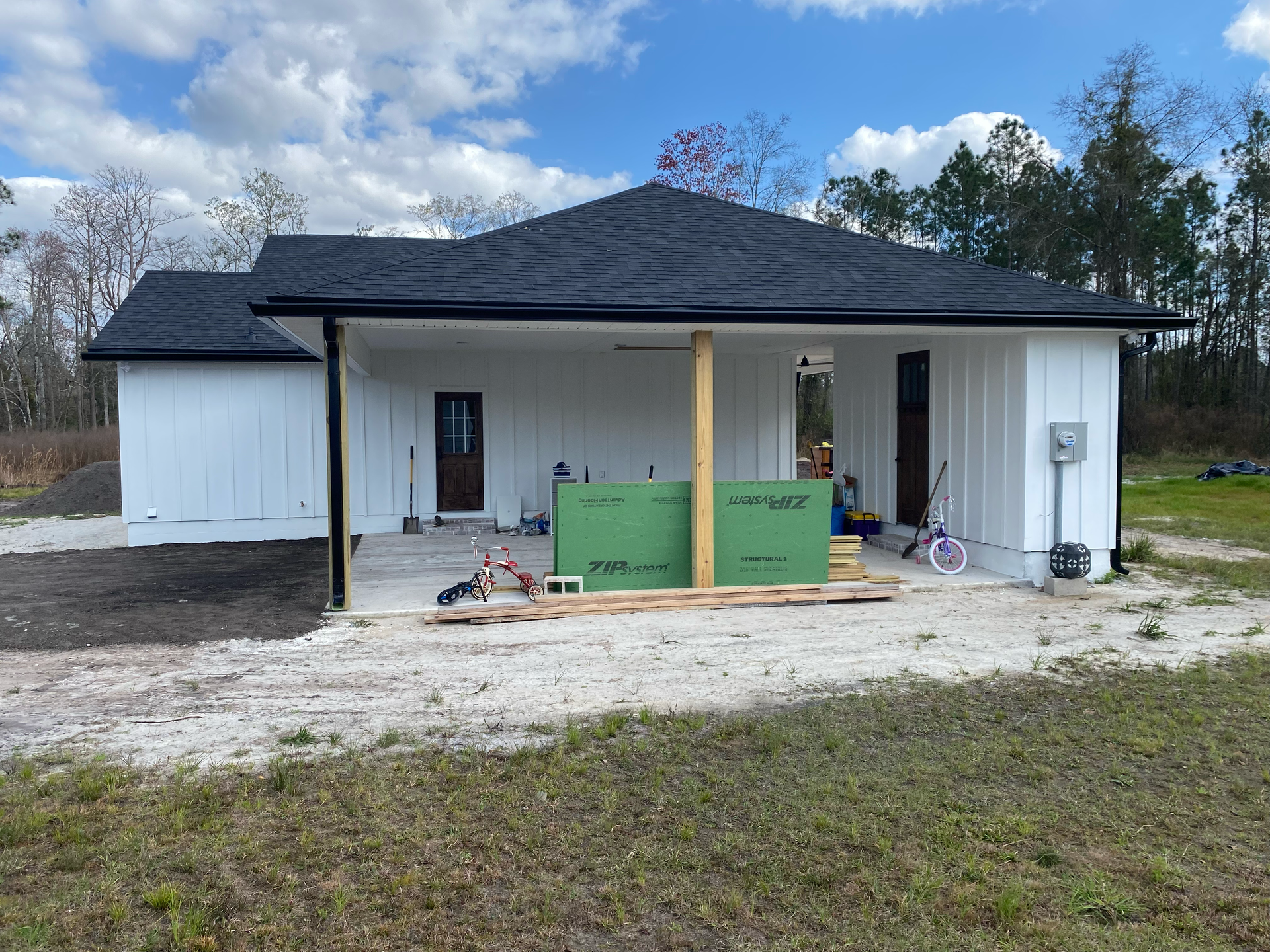 White house under construction with front porch, black roof, and green insulation panels in the yard