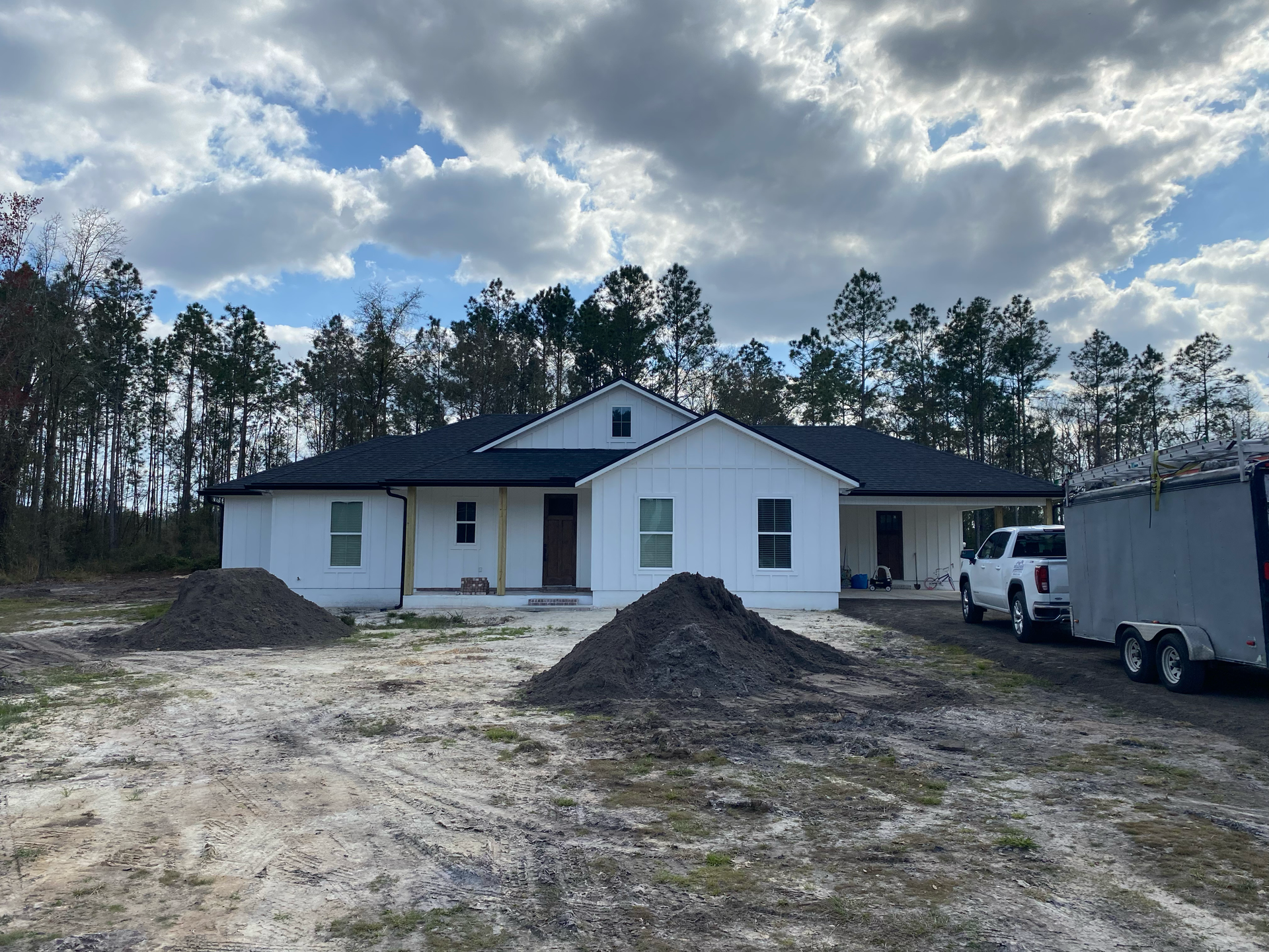 White single-story house under construction with dirt yard, piles of gravel, and a trailer parked beside it