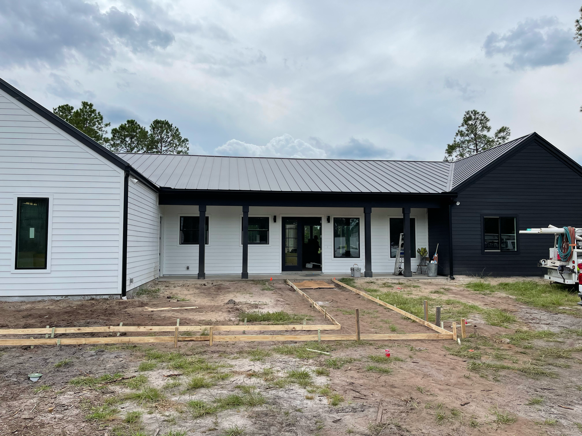Single-story white and black house exterior with porch, windows, and unfinished yard under cloudy sky