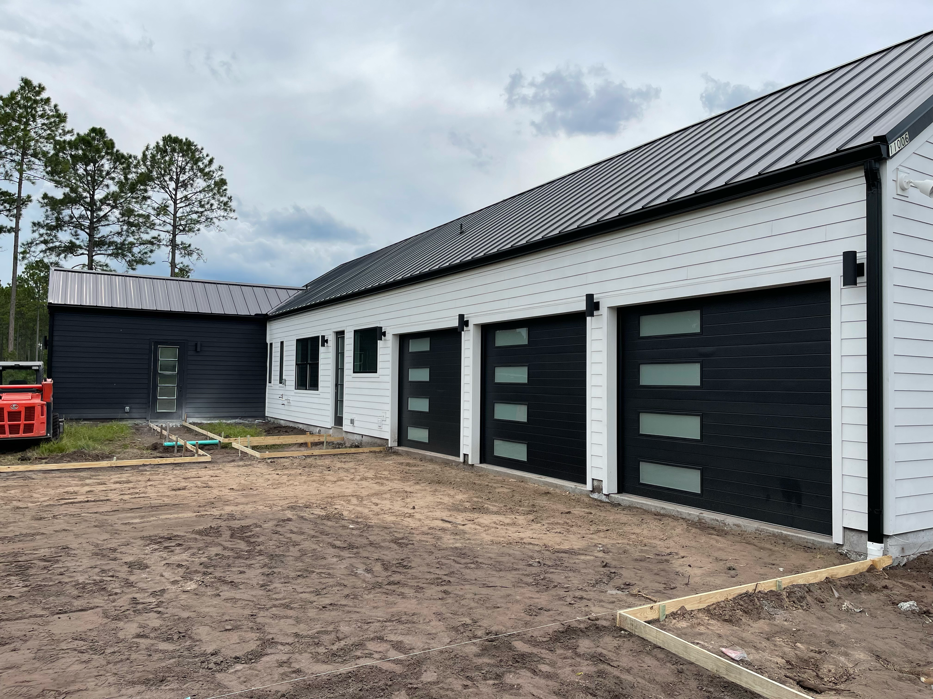 New white building with three black garage doors and a black-roofed outbuilding on a dirt lot