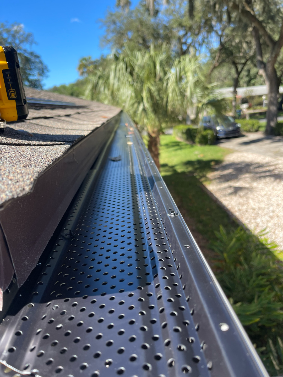 Close-up of a perforated metal roof gutter along a sloped roof beside trees and a sunny yard.