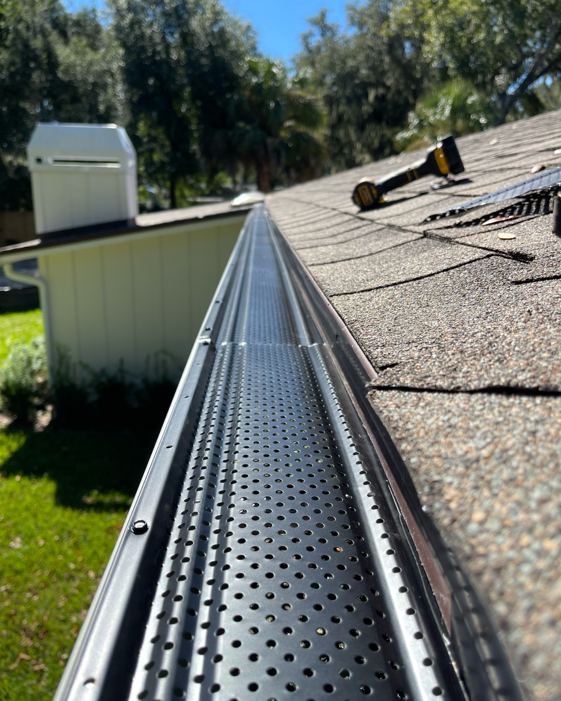 Perforated metal roof gutter along shingled rooftop, with trees and a shed in the background.