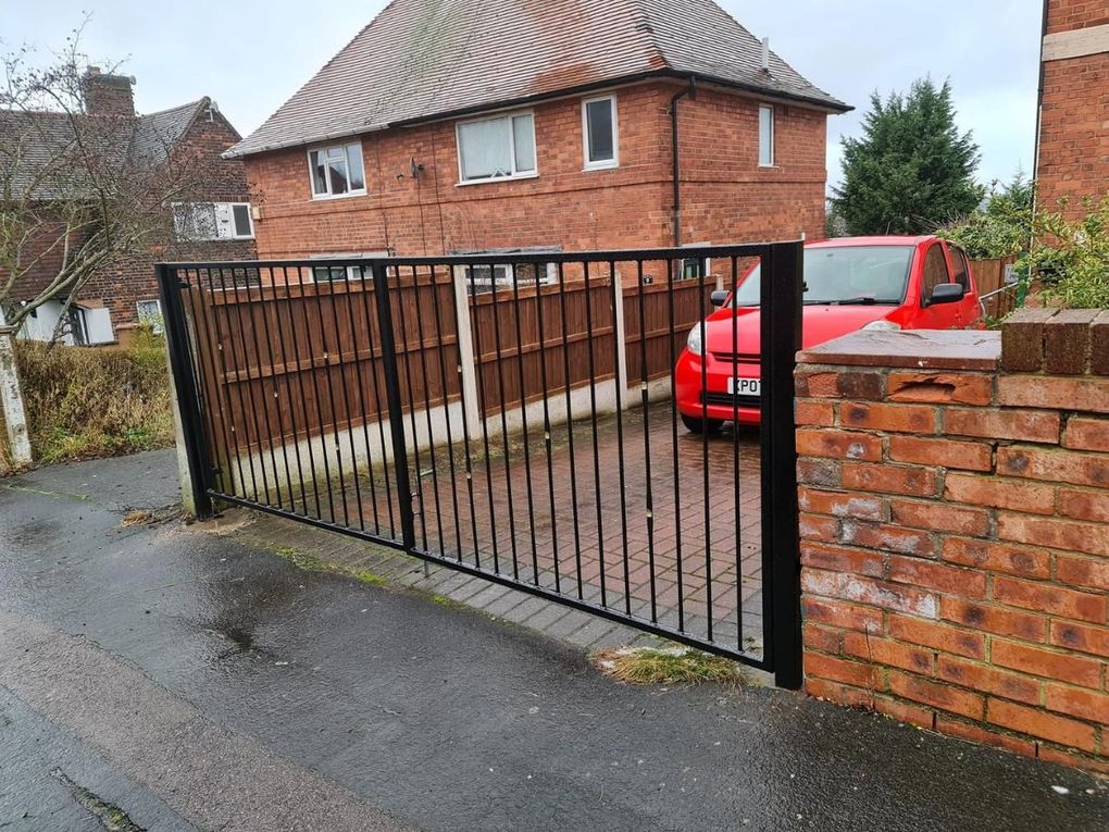Flat-topped black metal driveway gates with vertical bars, closing a block-paved drive beside a brick wall