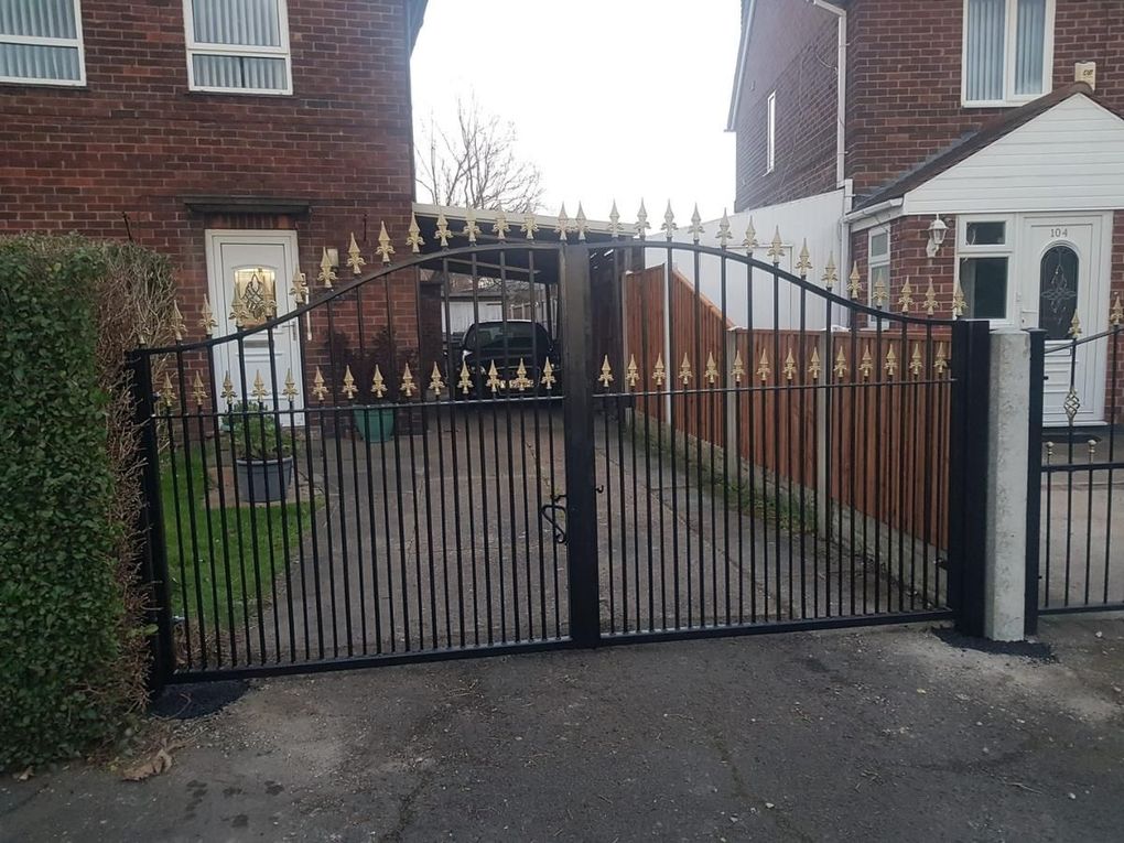 Arched black metal double driveway gates with two rows of gold fleur de lis finials, fitted across a block-paved drive