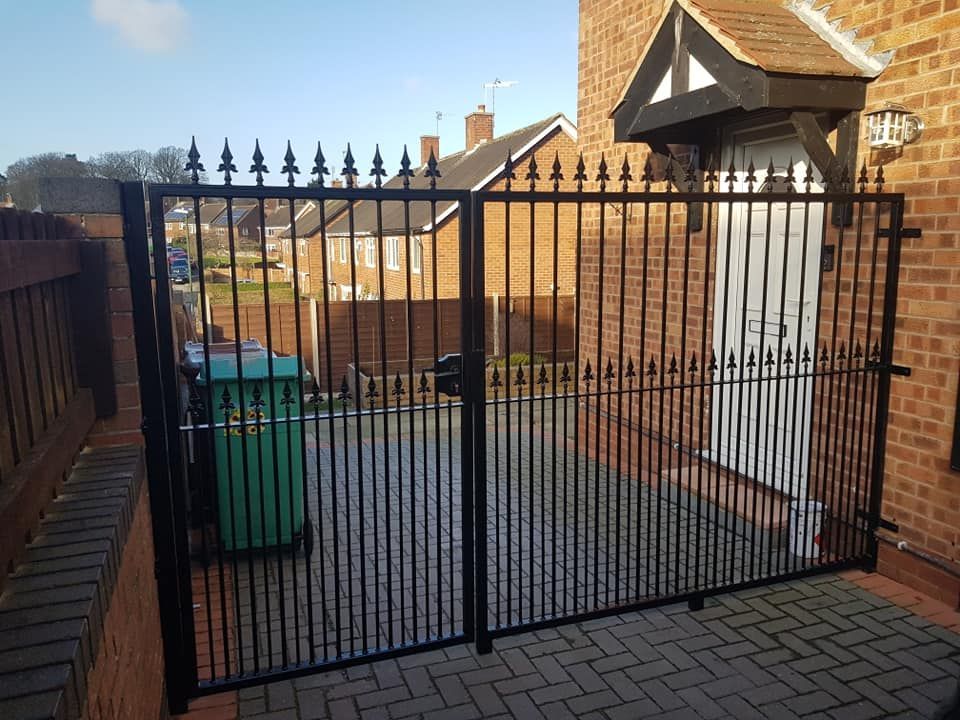 Black metal double driveway gates with fleur de lis heads set at two heights, across a herringbone block-paved drive