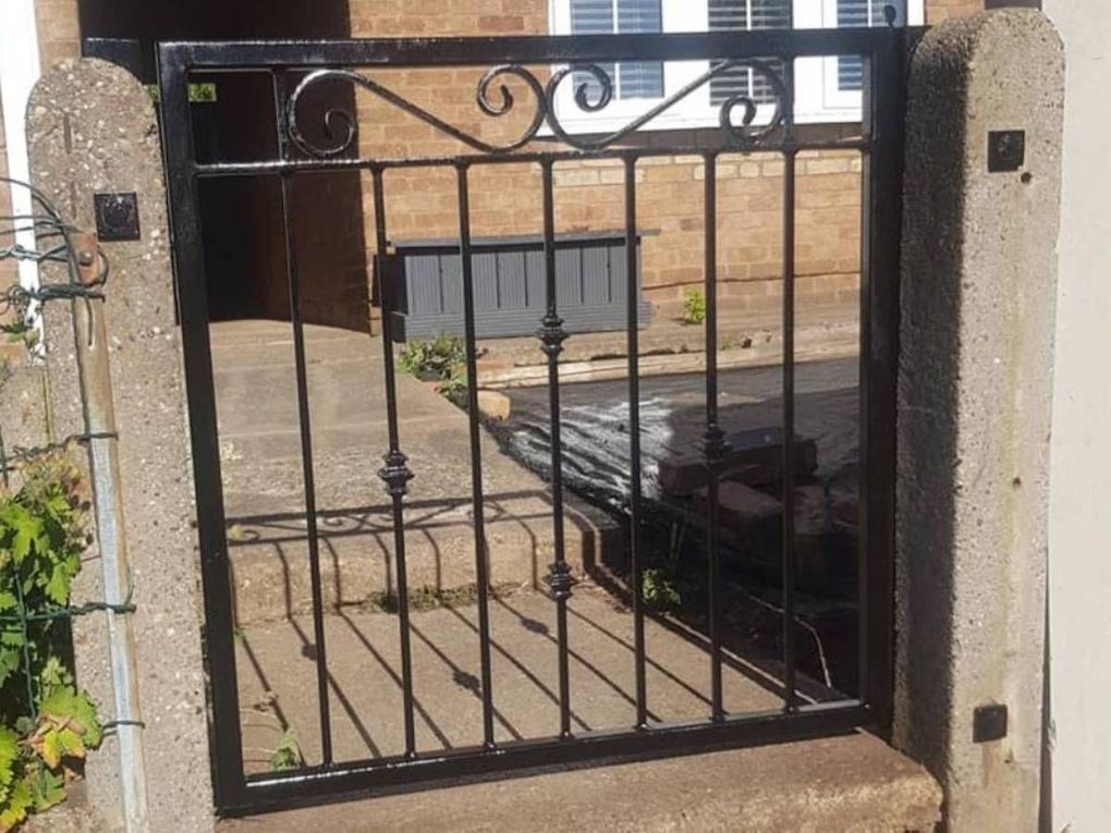 Black metal side gate with a scrolled top panel, hung between concrete posts