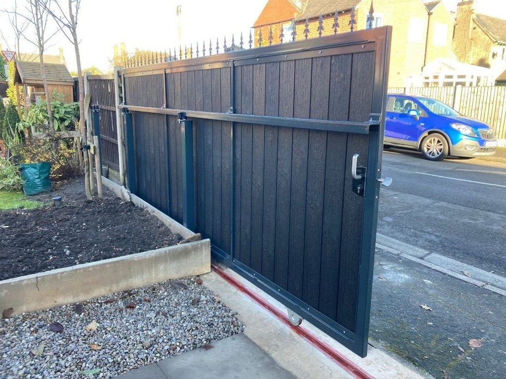 Black composite sliding gate with spear toppers across a roadside driveway, with the guide rail set in the ground