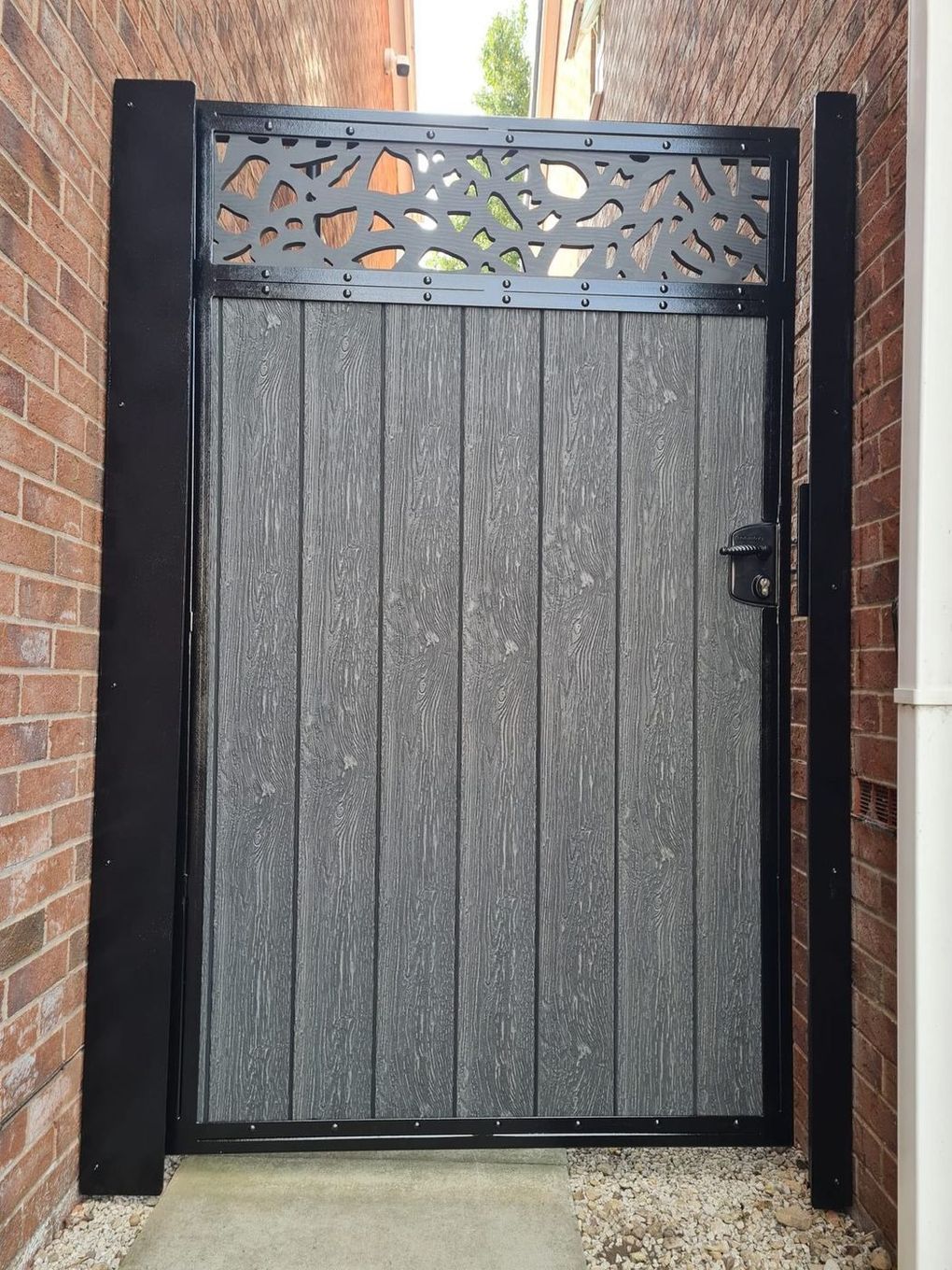 Graphite grey composite side gate with a laser-cut script-pattern top panel in a black frame, between two brick walls