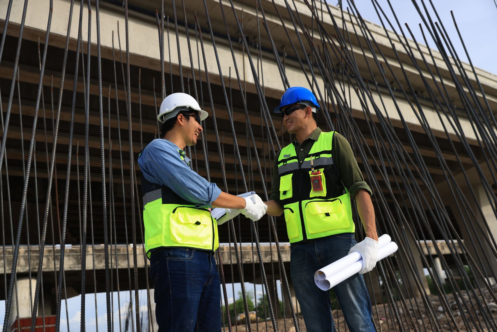 Two construction workers in safety vests and hard hats shake hands near rebar.