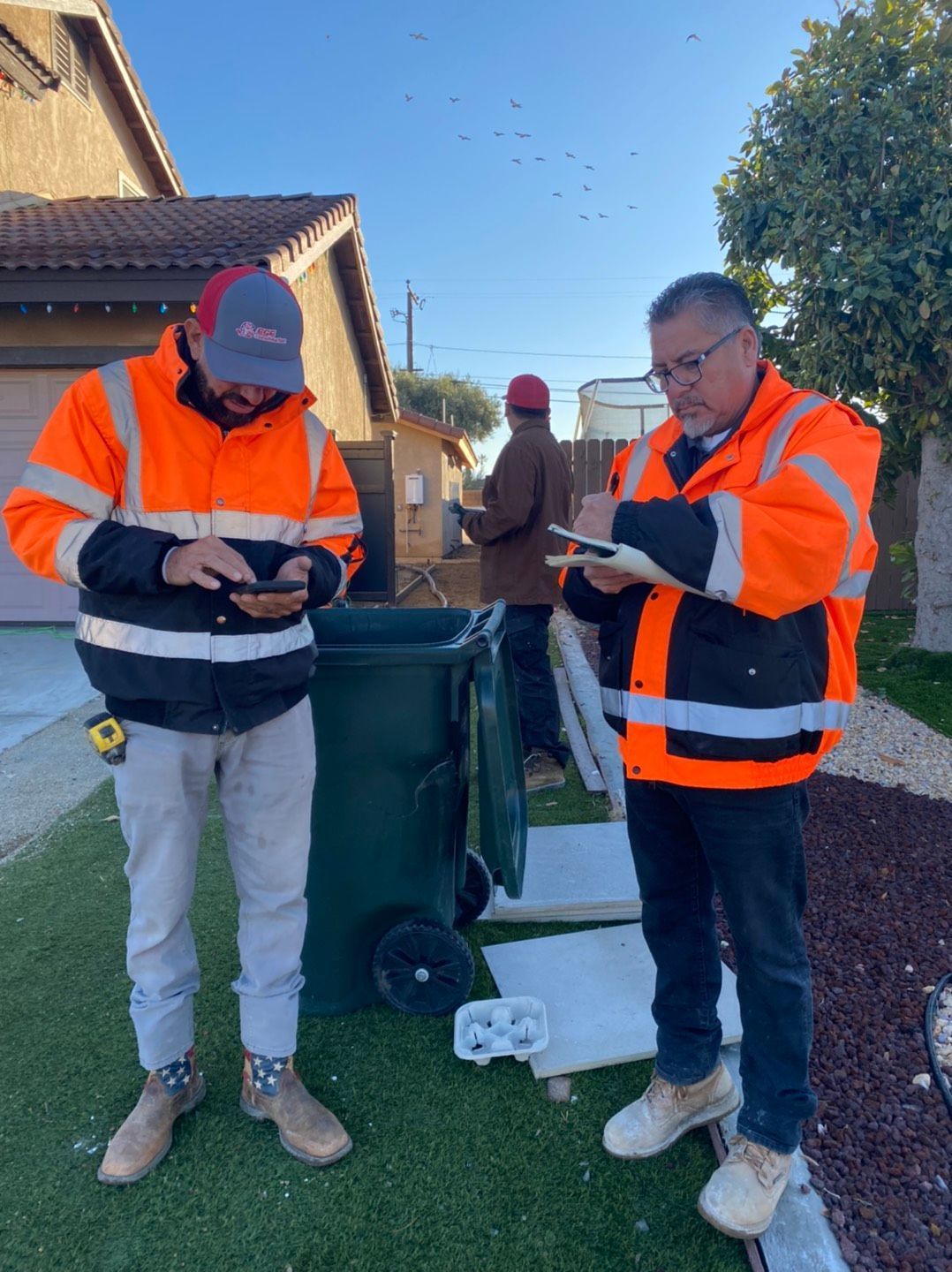 Three people in orange safety vests assess something near a house and trash can, one using a phone, one writing.