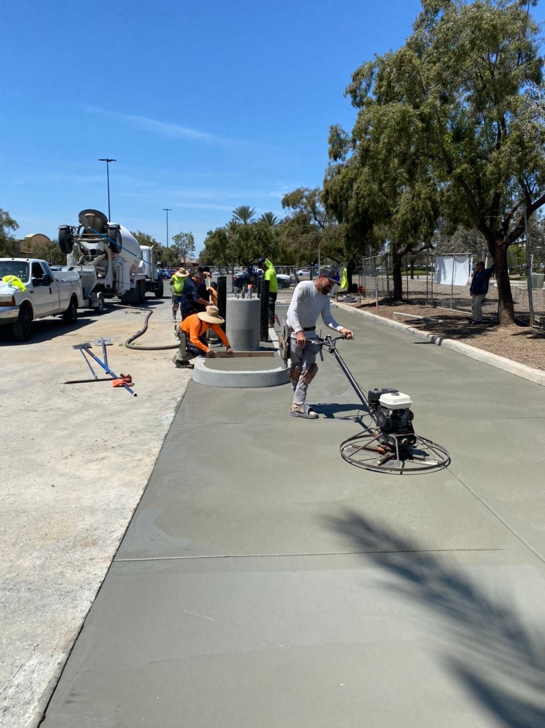 Construction workers smoothing wet concrete with a power trowel on a sunny day. A concrete truck is visible.