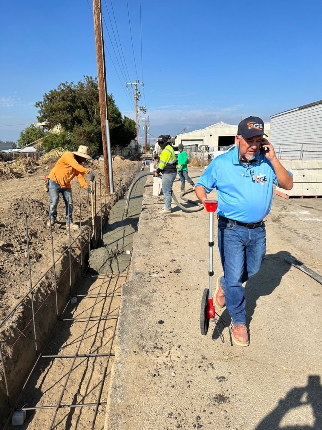 Construction workers building a wall outdoors. One talks on a phone while using a measuring wheel. Others pour concrete.