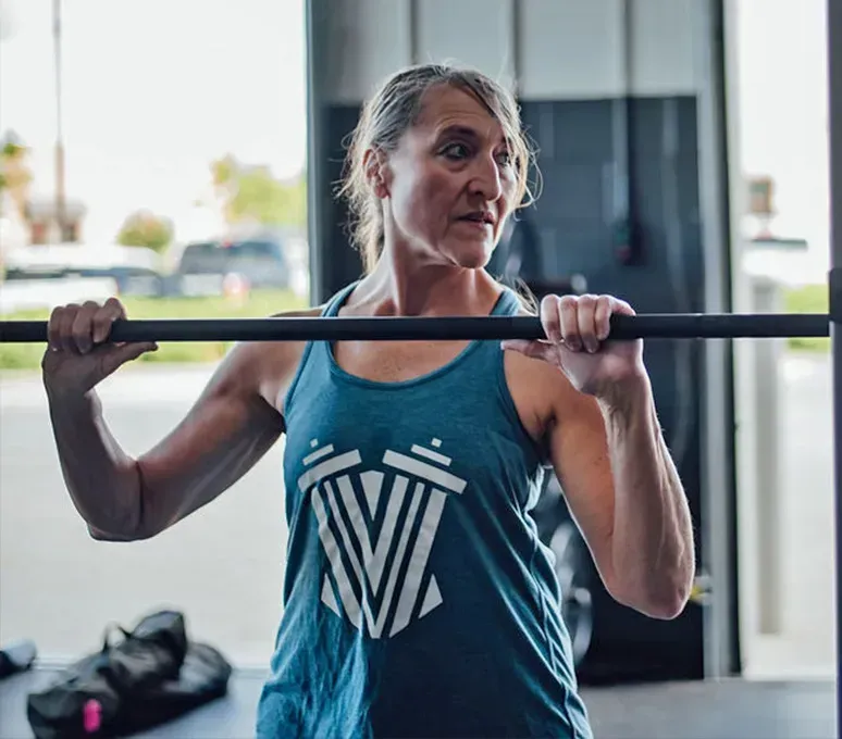 Woman holding barbell in gym, wearing a blue tank top, looking to her right.