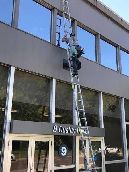A man on a ladder is cleaning the windows of a building that says 9 quality drive